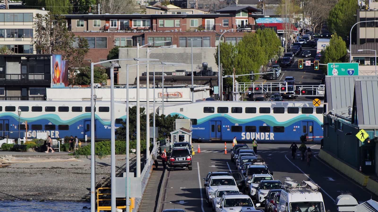 A Sounder commuter train crossing State Route 104 beside the Edmonds ferry terminal while cars queue on the ferry approach